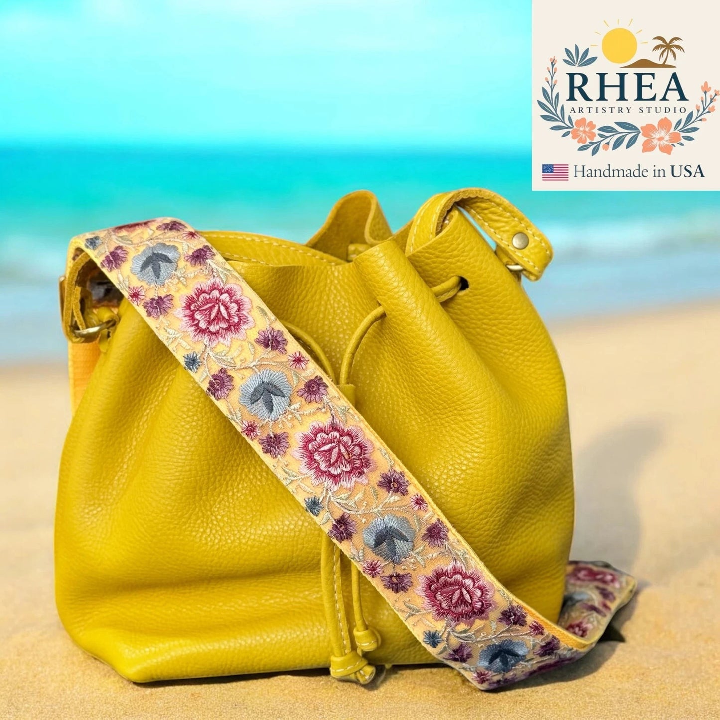 Rhea Artistry Studio 2-inch crossbody strap with floral embroidery in pink, blue, and yellow on a yellow leather bucket bag.