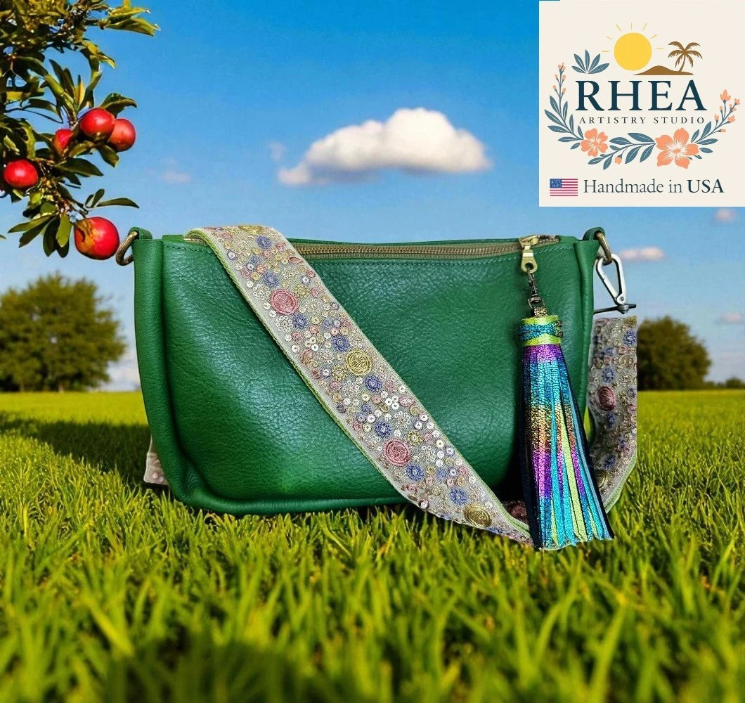 Rhea Artistry Studio green leather crossbody bag with a 2-inch wide embroidered strap featuring sun, moon, and floral motifs. Adjustable strap with brushed brass hardware and a colorful tassel.