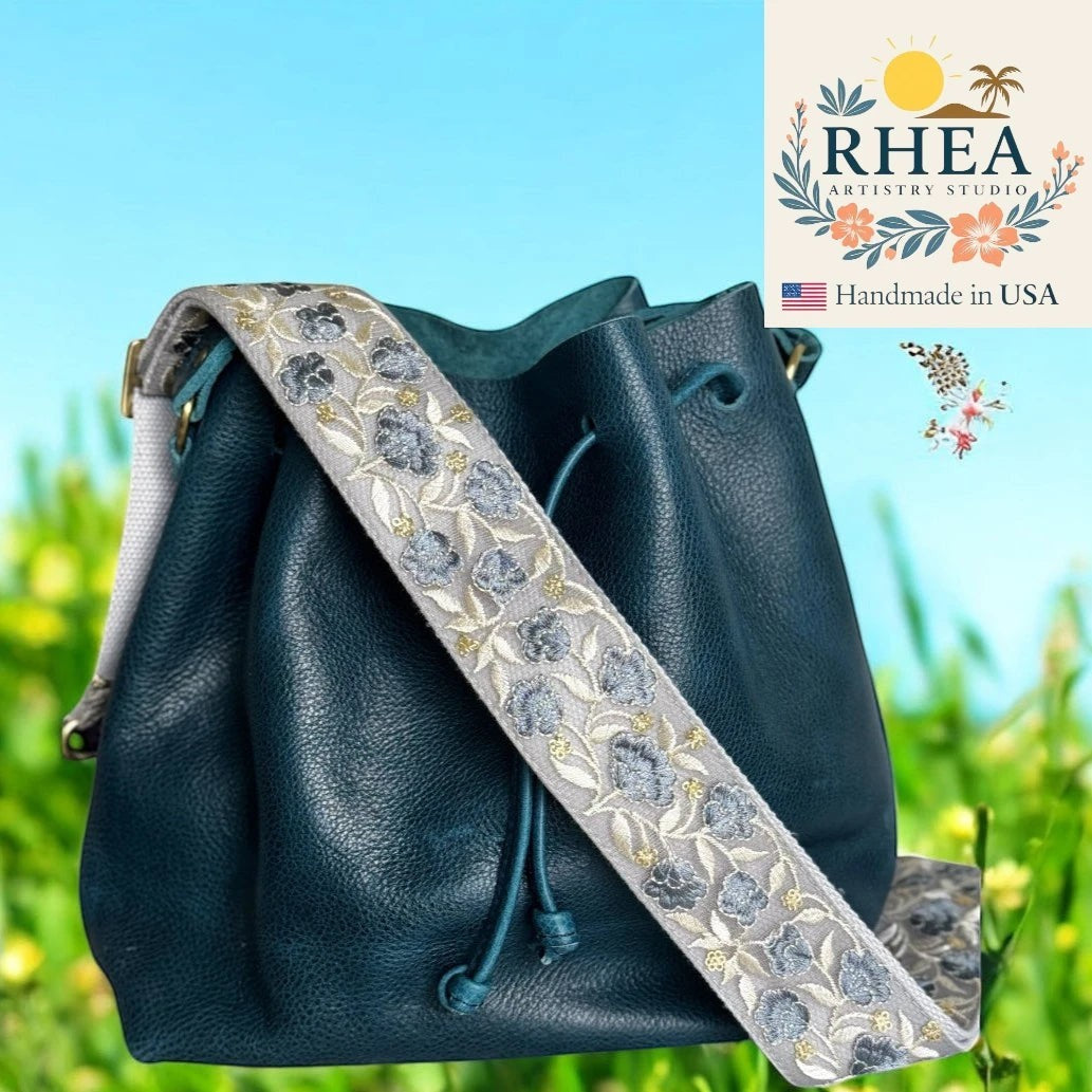 Rhea Artistry Studio 2-inch wide crossbody strap with dusty blue floral embroidery on a light grey background, featuring brushed brass hardware.