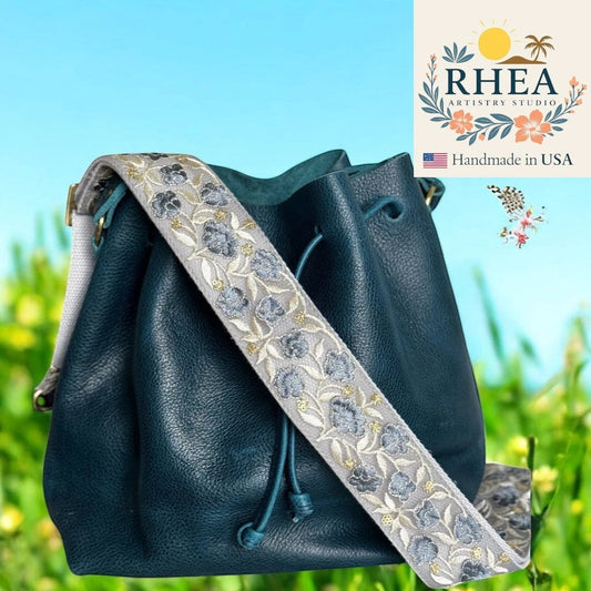Rhea Artistry Studio 2-inch wide crossbody strap with dusty blue floral embroidery on a light grey background, featuring brushed brass hardware.