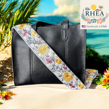 Rhea Artistry Studio grey crossbody strap with yellow, pink, and white floral embroidery and brushed brass hardware.