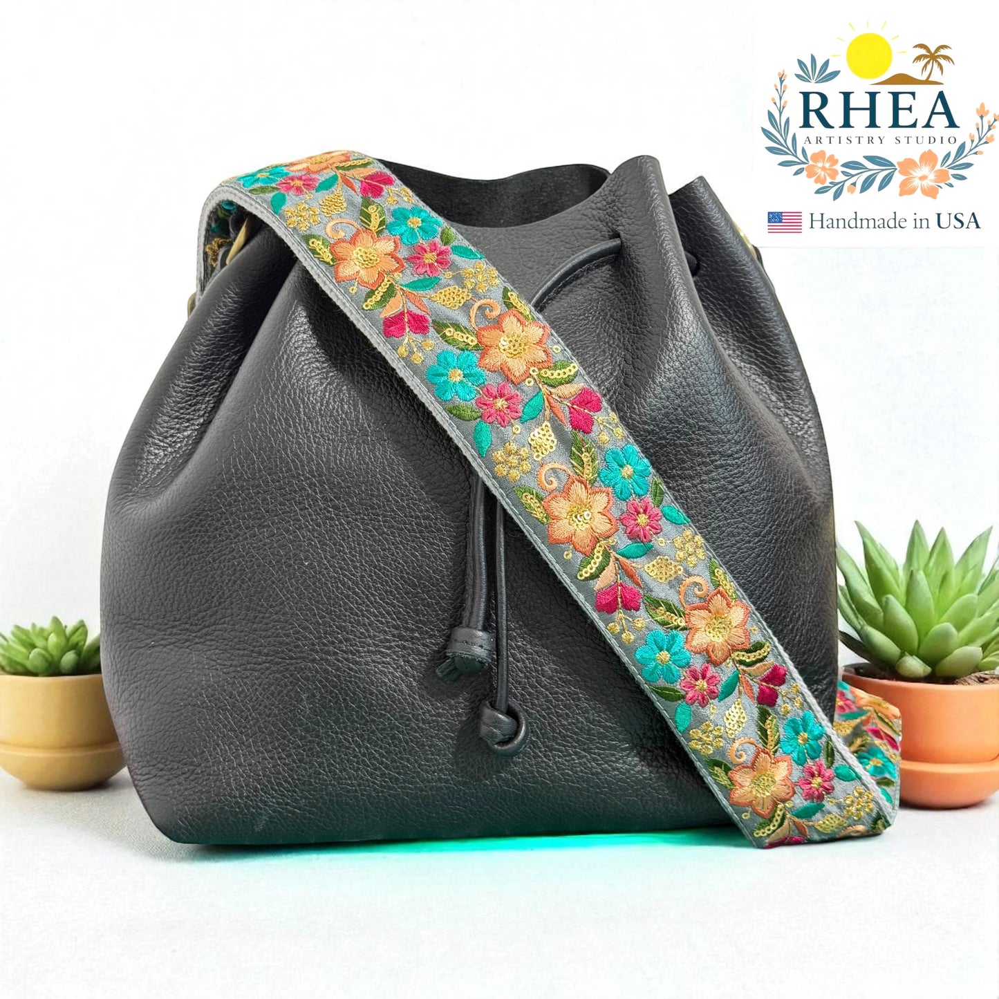 Rhea Artistry Studio 2-inch wide grey crossbody strap with vibrant floral embroidery in pink, orange, and teal. Adjustable, cotton webbing backing, brushed brass hardware.