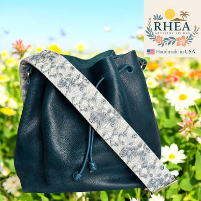 Rhea Artistry Studio 2-inch wide crossbody strap with embroidered frosted flora pattern in grey and white on a teal leather bucket bag.
