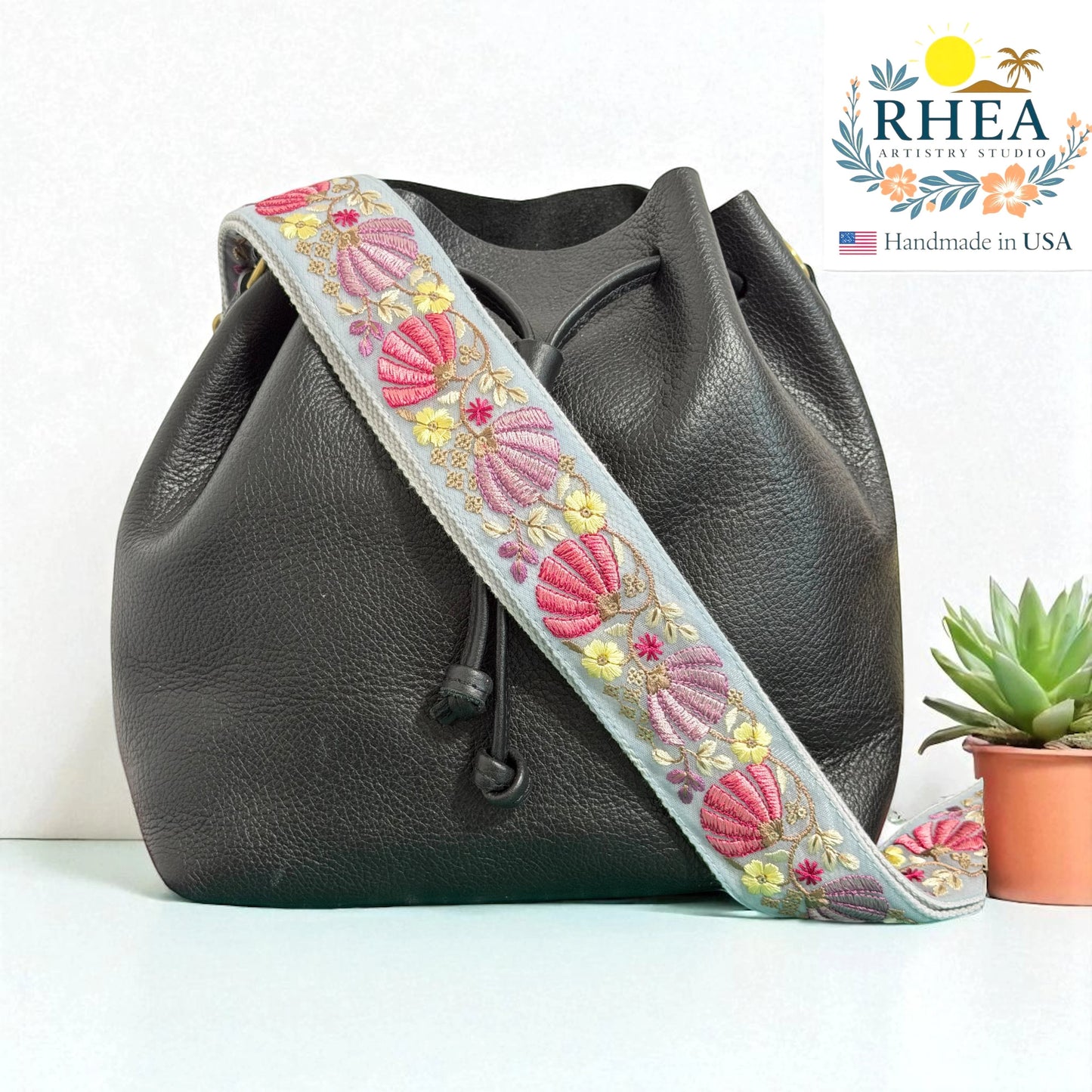 Rhea Artistry Studio light blue embroidered crossbody strap with pink and yellow floral details on a black leather bucket bag.