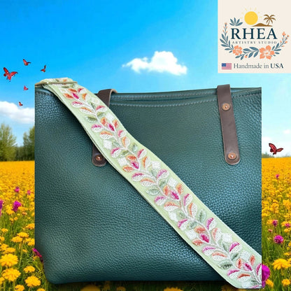 Rhea Artistry Studio light green embroidered crossbody strap with floral leaf pattern and brushed brass hardware on a dark green pebbled leather tote bag.