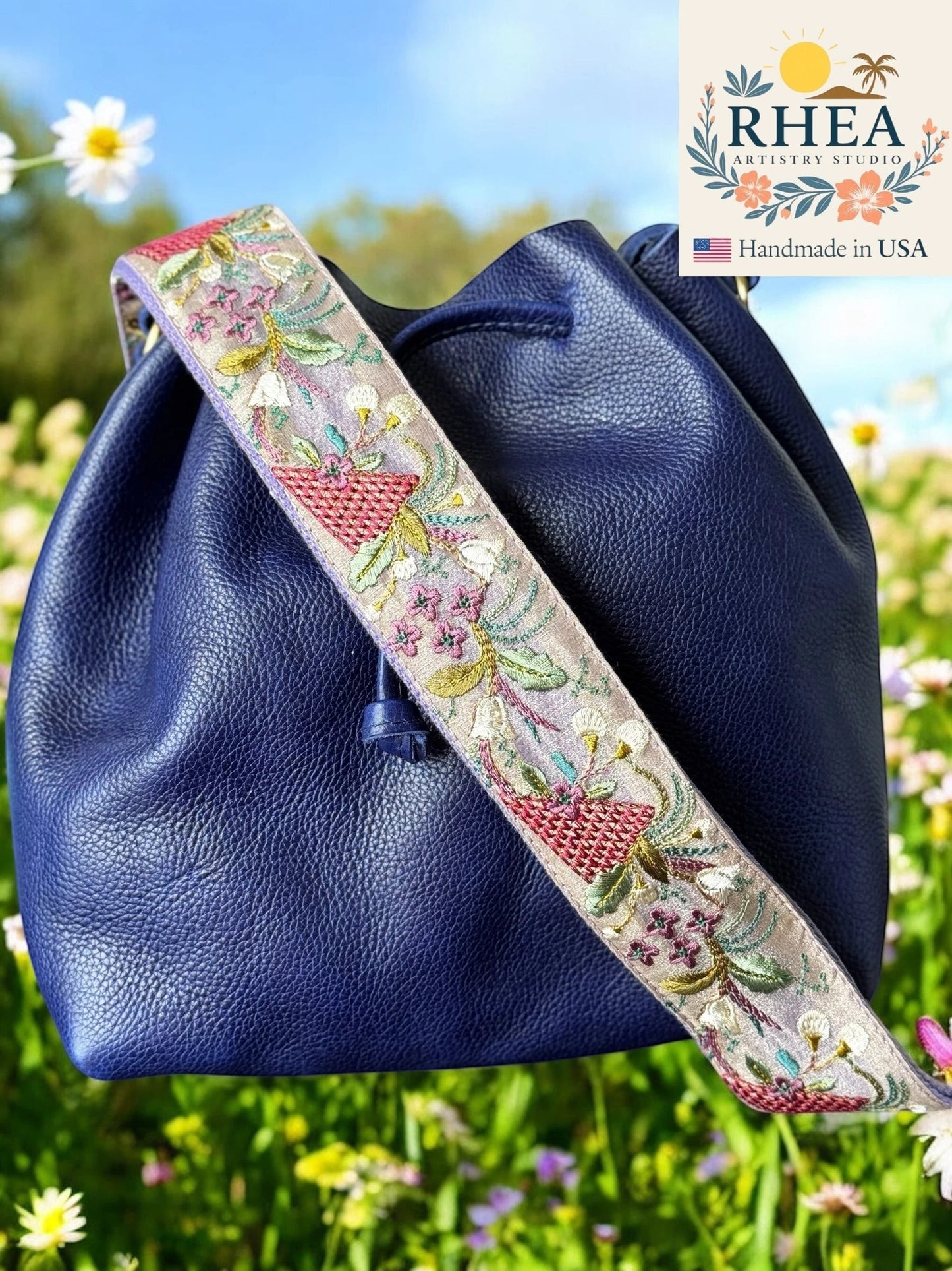 Rhea Artistry Studio navy blue leather bucket bag with a 2-inch wide embroidered strap featuring floral and wheat field motifs in light taupe, pink, and green.