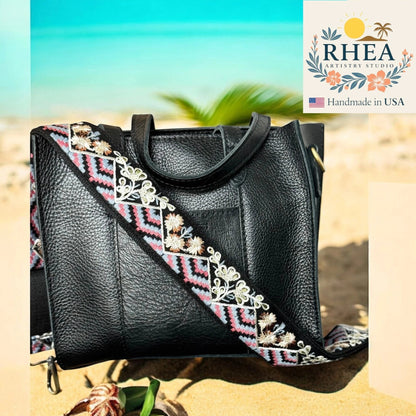 Rhea Artistry Studio black leather crossbody bag with a 2-inch wide adjustable embroidered strap featuring floral and geometric patterns in pink, blue, and white.
