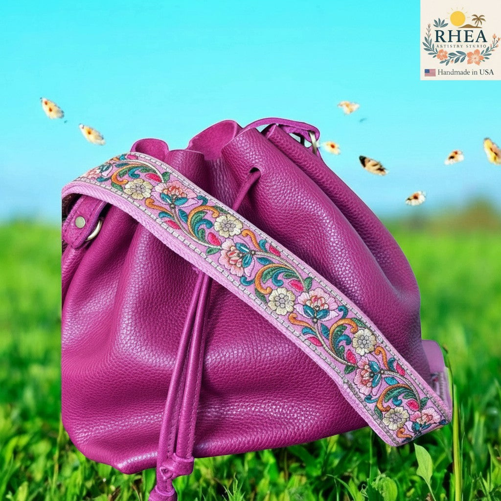 Rhea Artistry Studio 2-inch wide crossbody strap with colorful floral embroidery on pink lace, attached to a magenta leather bucket bag.