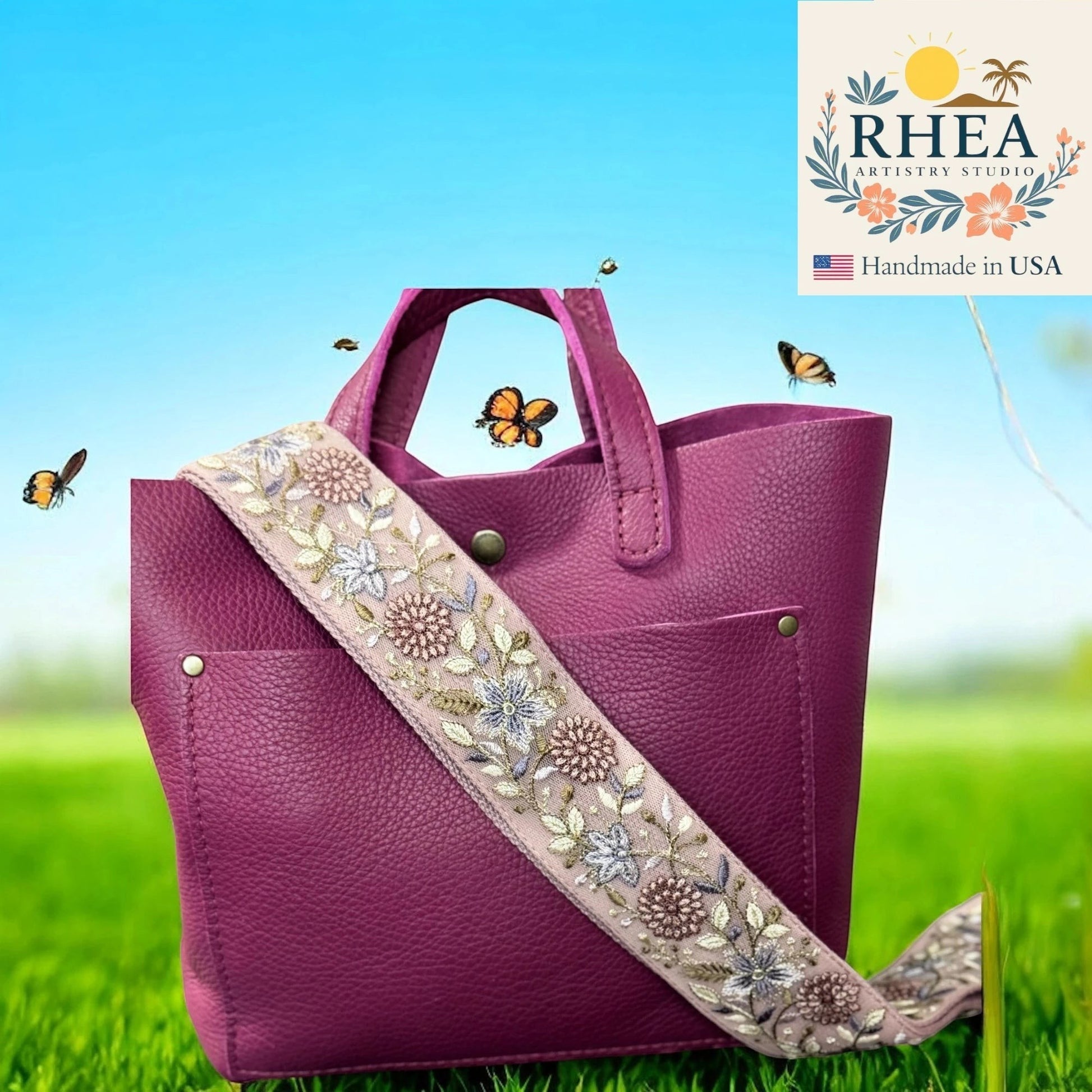 Rhea Artistry Studio 2-inch wide crossbody strap with floral embroidery in pink, blue, and silver on a light pink background. Adjustable strap with brass hardware.