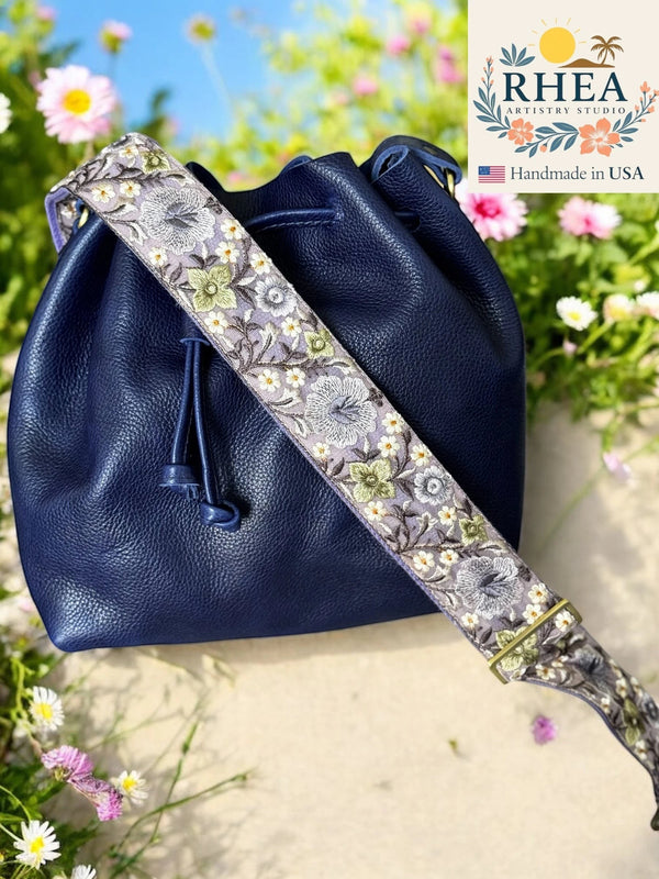 Rhea Artistry Studio 2-inch wide crossbody strap with detailed floral embroidery in purple, green, and white on a light grey background, attached to a navy blue leather bucket bag.