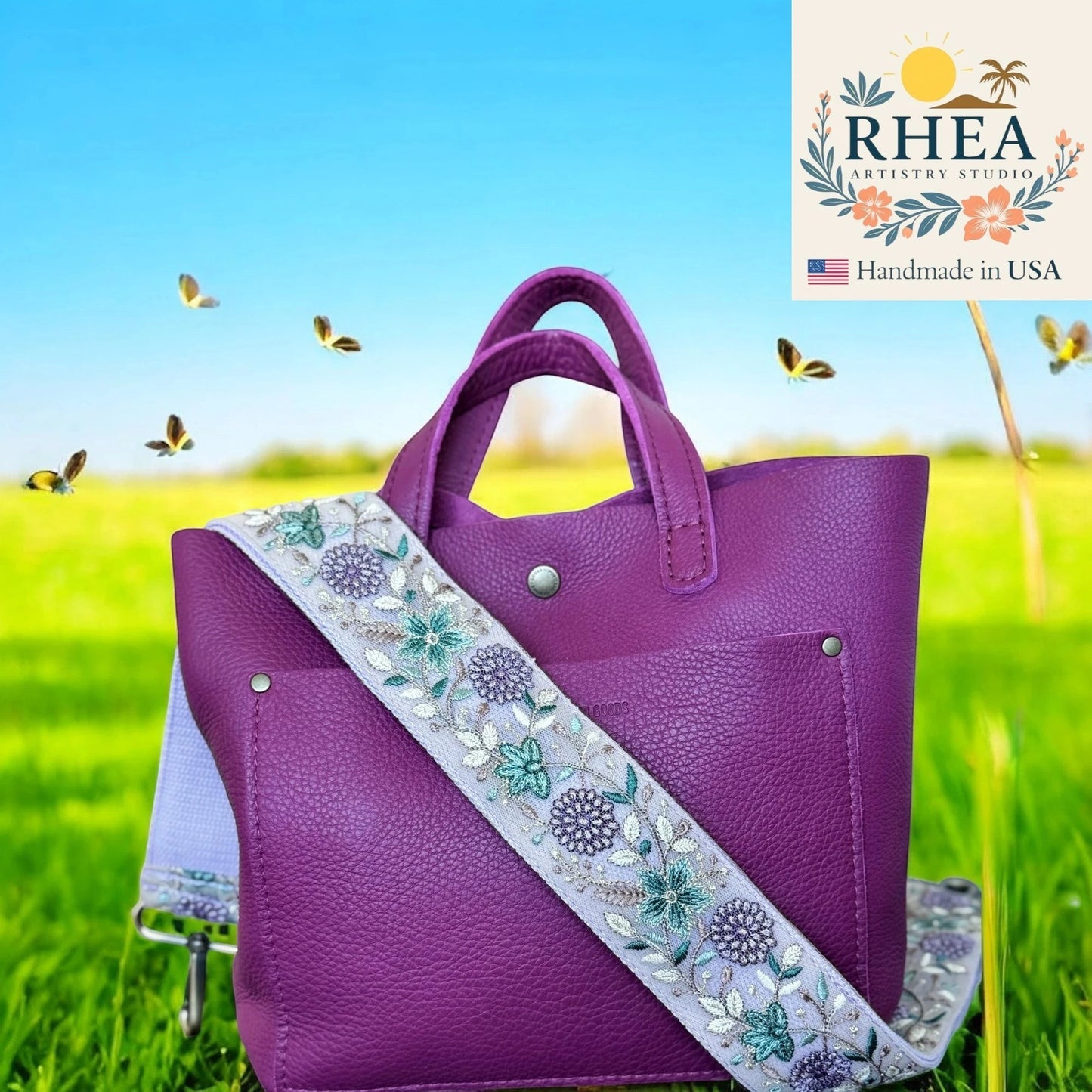Rhea Artistry Studio 2-inch wide crossbody strap with floral embroidery in purple, green, and white on a light grey background. Adjustable strap with brushed brass hardware.