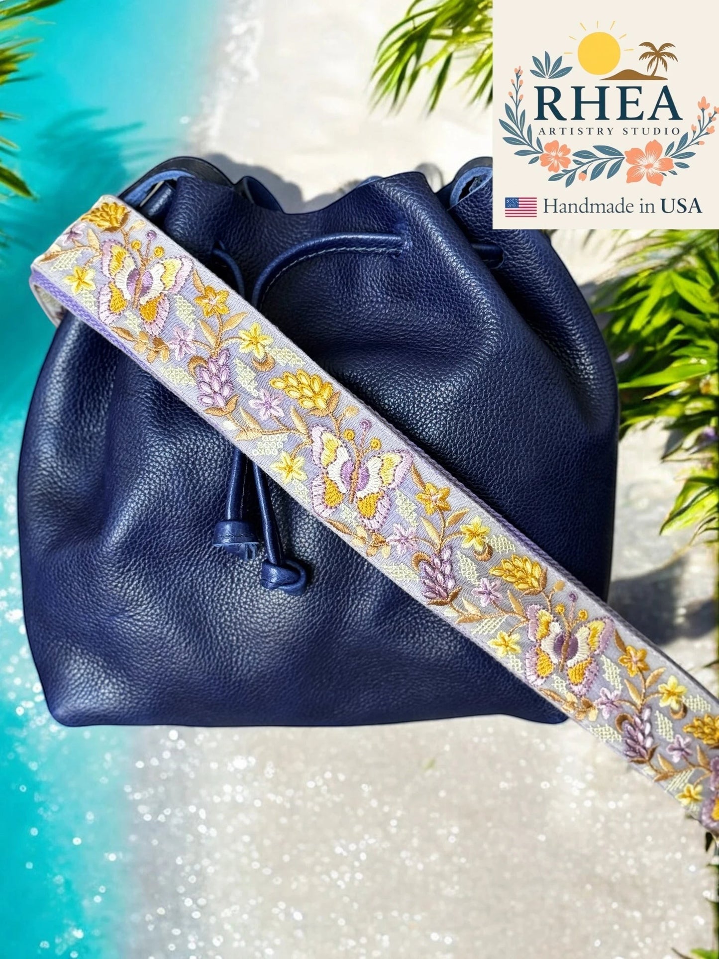 Rhea Artistry Studio navy blue leather bucket bag with a 2-inch wide adjustable crossbody strap featuring intricate purple and yellow floral embroidery.