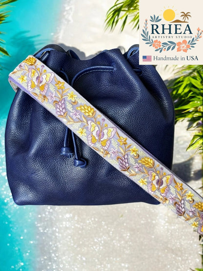 Rhea Artistry Studio navy blue leather bucket bag with a 2-inch wide adjustable crossbody strap featuring intricate purple and yellow floral embroidery.