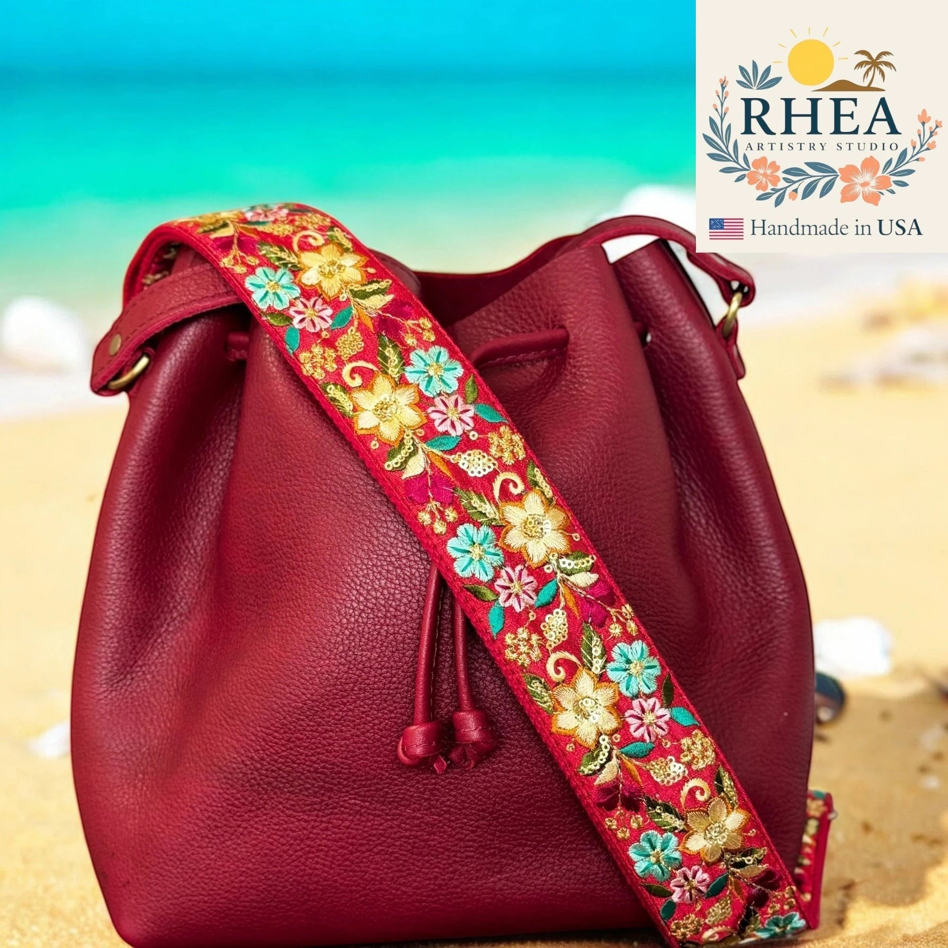 Rhea Artistry Studio 2-inch wide adjustable crossbody strap with vibrant floral embroidery on a red background, attached to a burgundy leather bucket bag on a beach.
