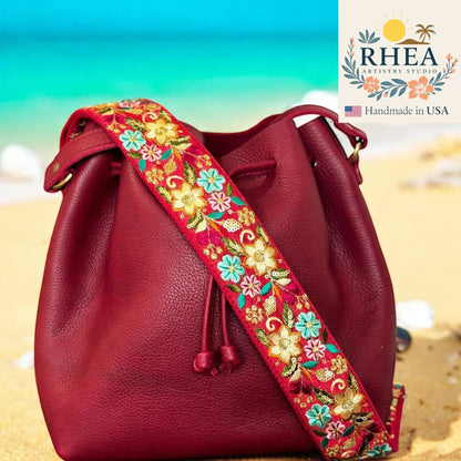 Rhea Artistry Studio 2-inch wide adjustable crossbody strap with vibrant floral embroidery on a red background, attached to a burgundy leather bucket bag on a beach.