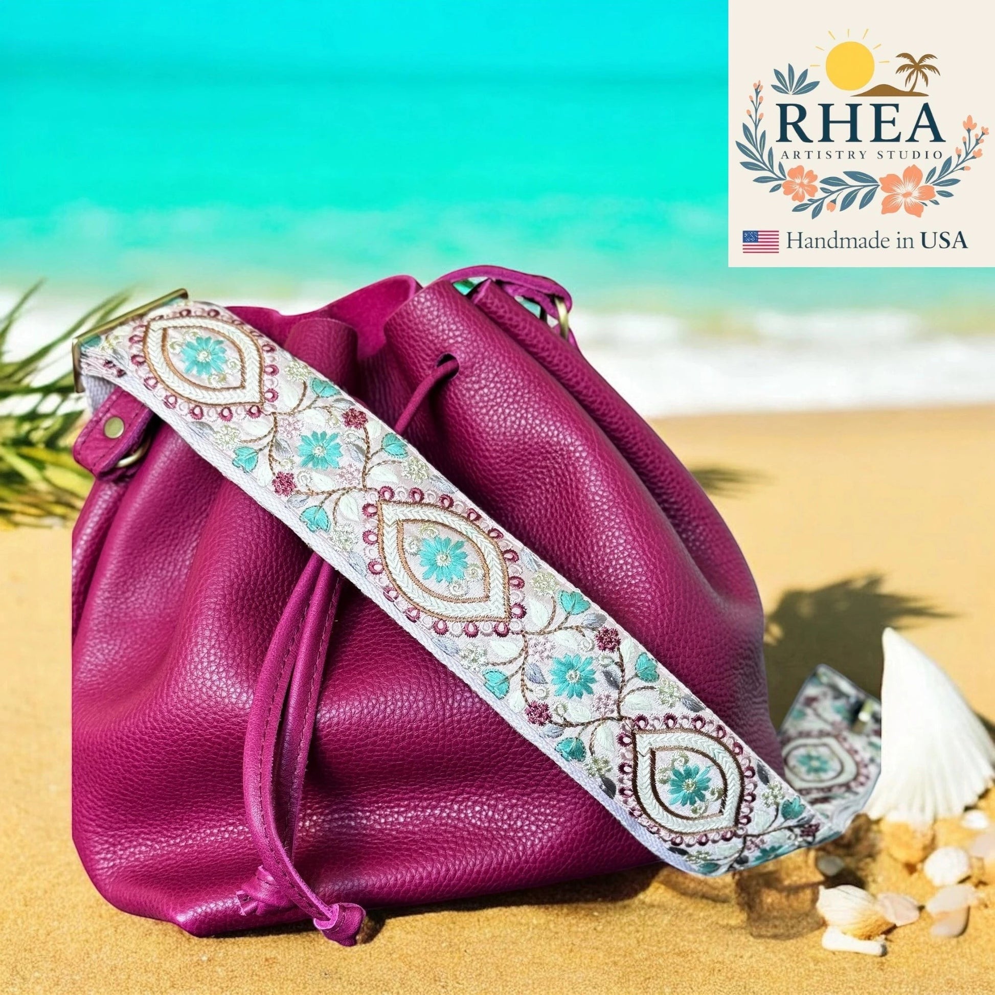 Rhea Artistry Studio 2-inch crossbody strap with embroidered floral panels in teal and burgundy on a white background, attached to a magenta leather bucket bag on a beach.