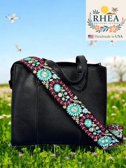 Rhea Artistry Studio 2-inch wide crossbody strap with dahlia floral embroidery in teal, pink, and white on black webbing. Adjustable length with brushed brass hardware.
