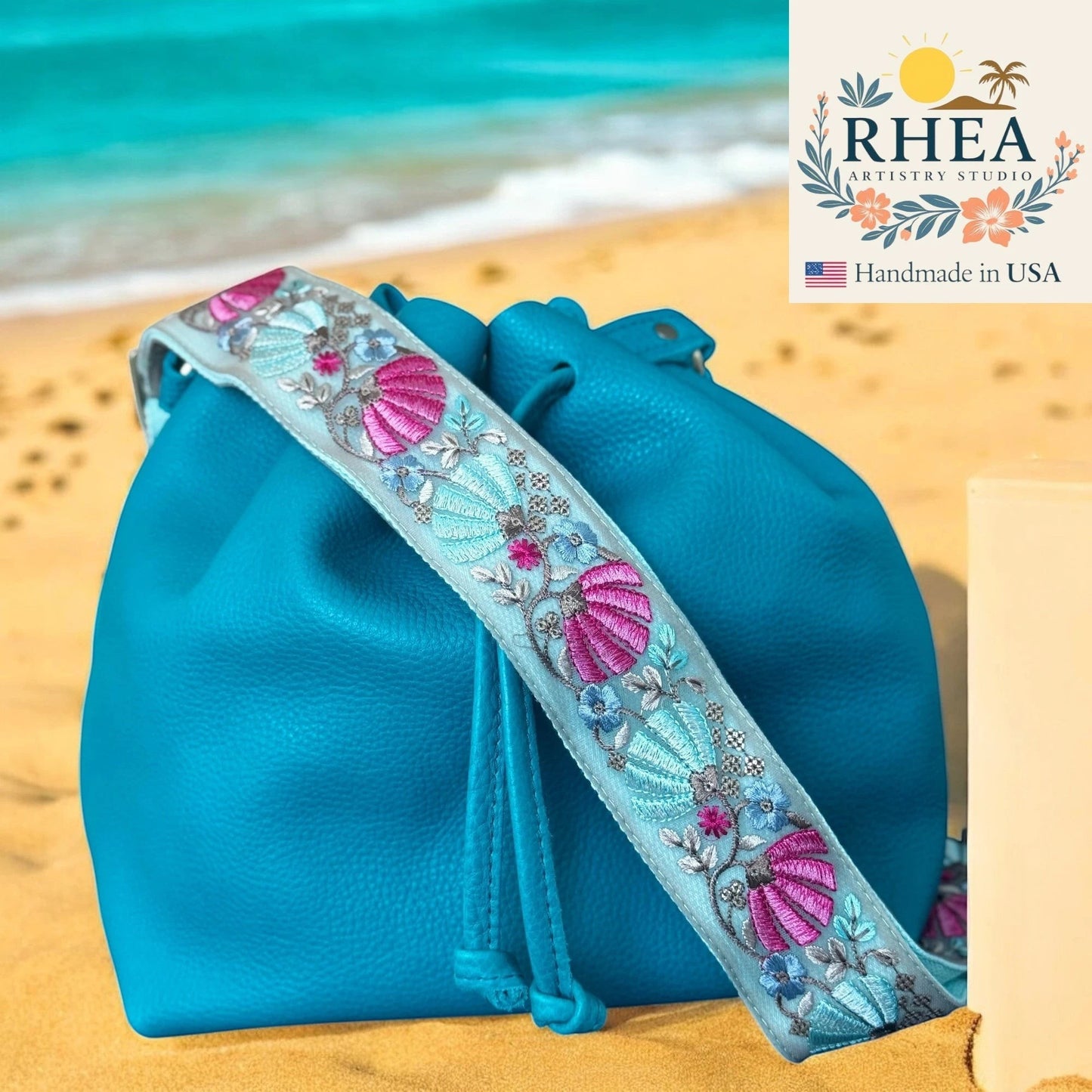 Rhea Artistry Studio light blue embroidered crossbody strap with pink and teal floral and shell motifs, attached to a teal leather bucket bag on a beach.