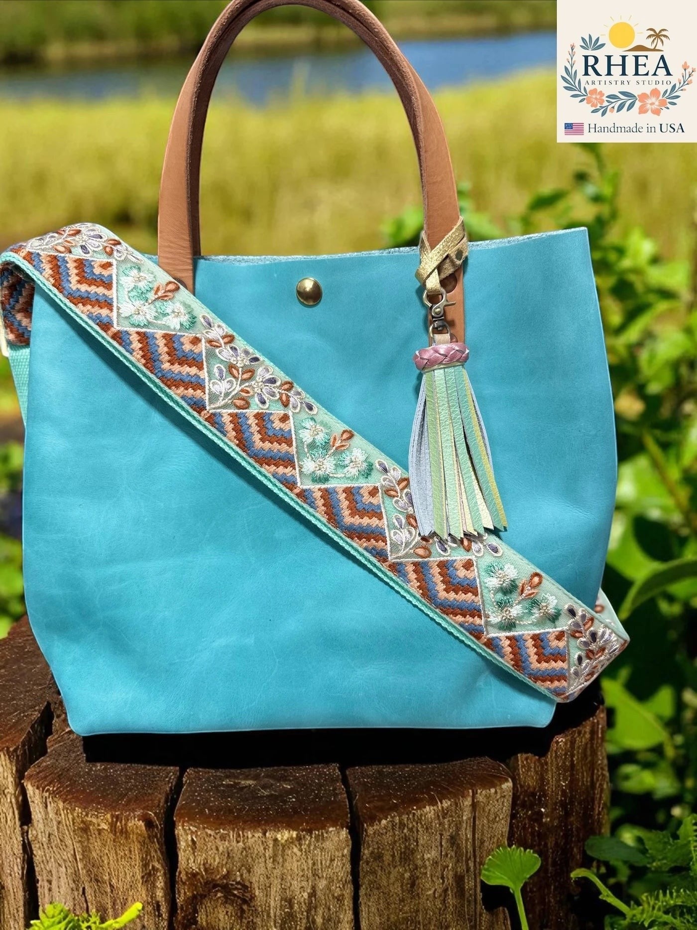Rhea Artistry Studio turquoise leather tote bag with a 2-inch wide adjustable embroidered strap featuring floral and geometric patterns in blue, brown, and green.