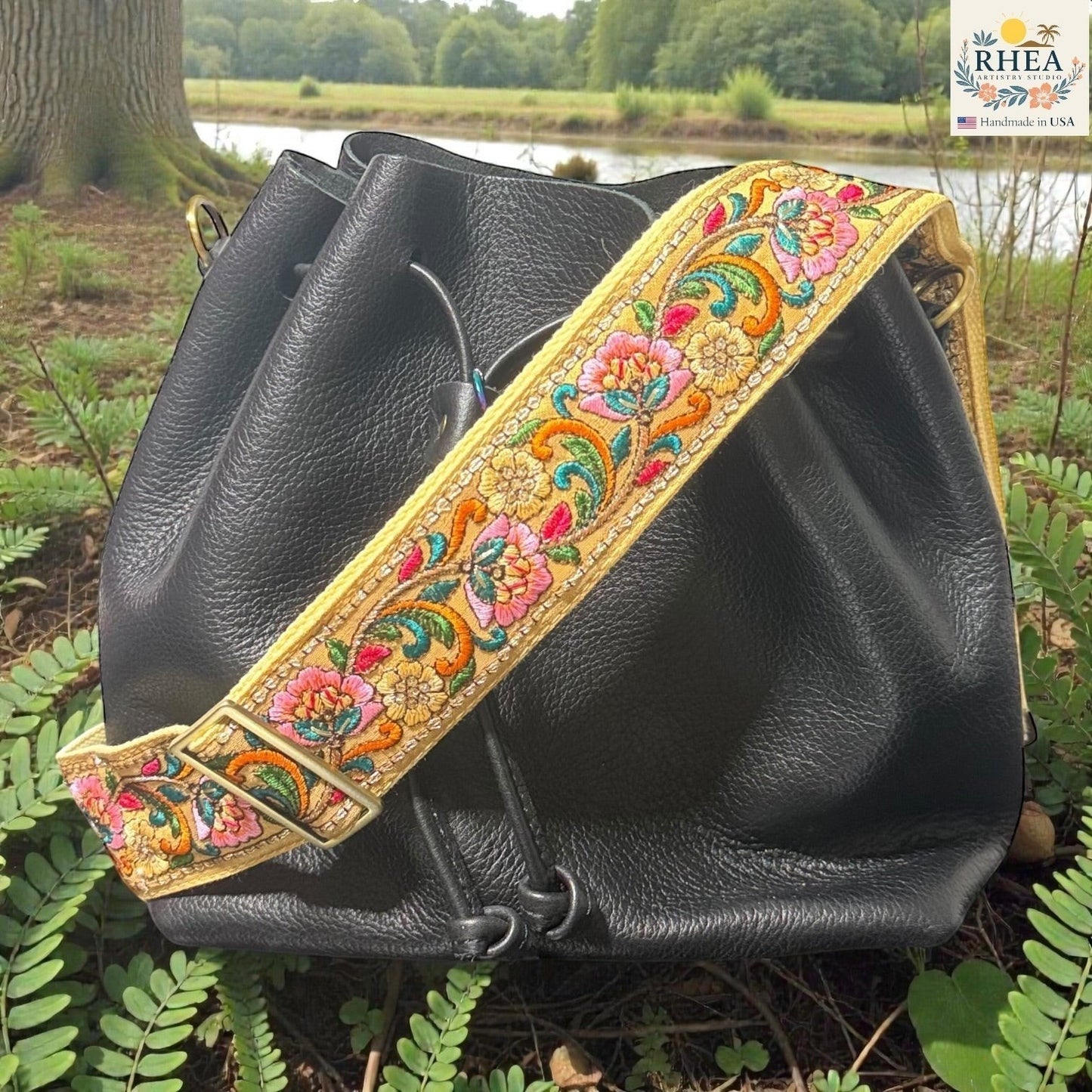 Rhea Artistry Studio yellow crossbody strap with colorful floral embroidery and brushed brass hardware, shown on a black leather bucket bag.