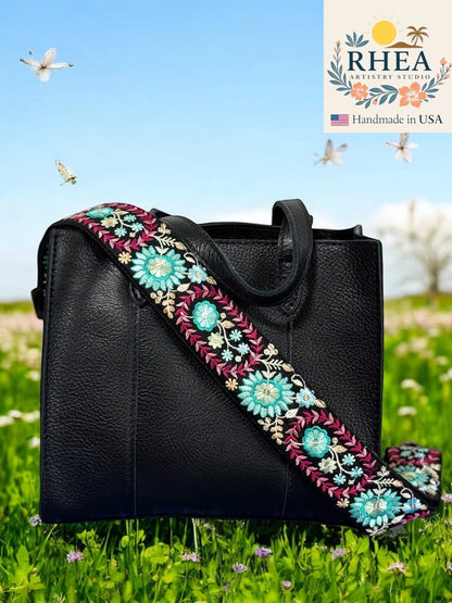 Rhea Artistry Studio 2-inch wide crossbody strap with dahlia floral embroidery in teal, pink, and white on black webbing. Adjustable length with brass hardware.