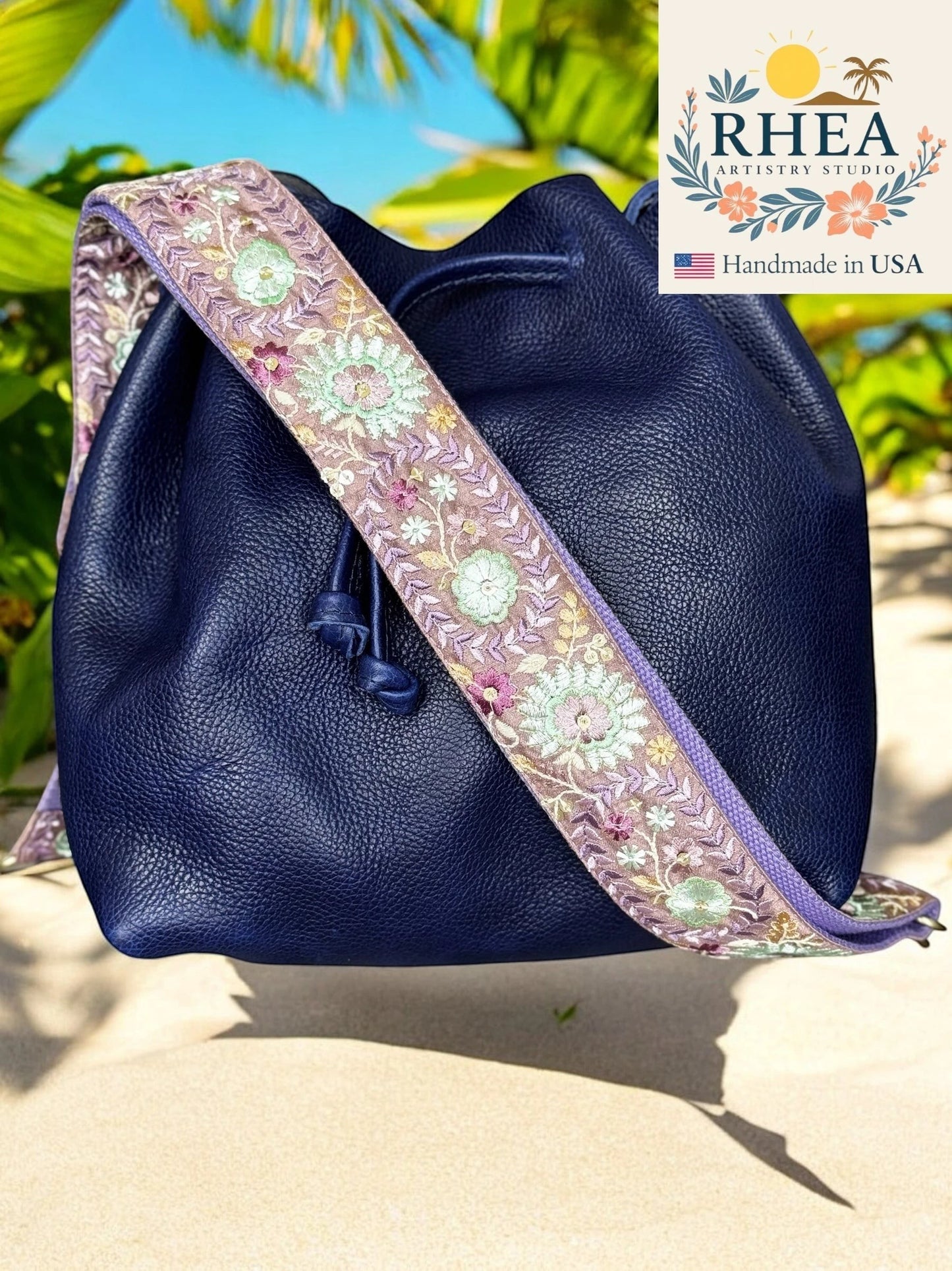 Rhea Artistry Studio navy blue leather bucket bag with a 2-inch wide adjustable embroidered strap featuring dahlia floral patterns in purple, green, and pink.