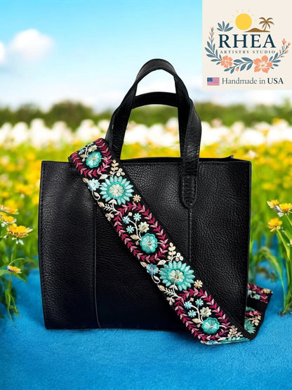 Rhea Artistry Studio 2-inch wide crossbody strap with dahlia floral embroidery in teal, pink, and blue on black webbing. Adjustable length with brushed brass hardware.