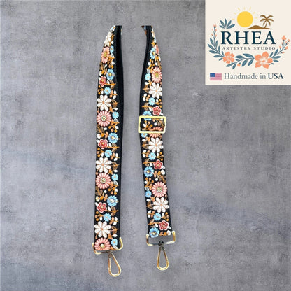 Rhea Artistry Studio black crossbody strap with embroidered colorful daisy flowers, adjustable length, and brushed brass hardware.
