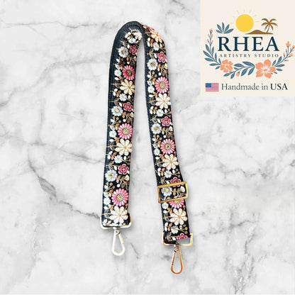 Rhea Artistry Studio black crossbody strap with embroidered pink, white, and blue daisy flowers, adjustable length, brushed brass hardware.