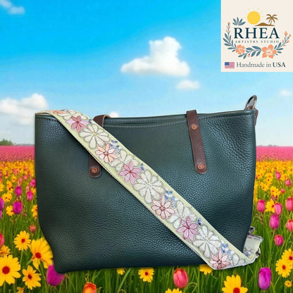 Rhea Artistry Studio 2-inch crossbody strap with embroidered daisies in pink, white, and blue on a light green background. Adjustable strap with brass hardware.