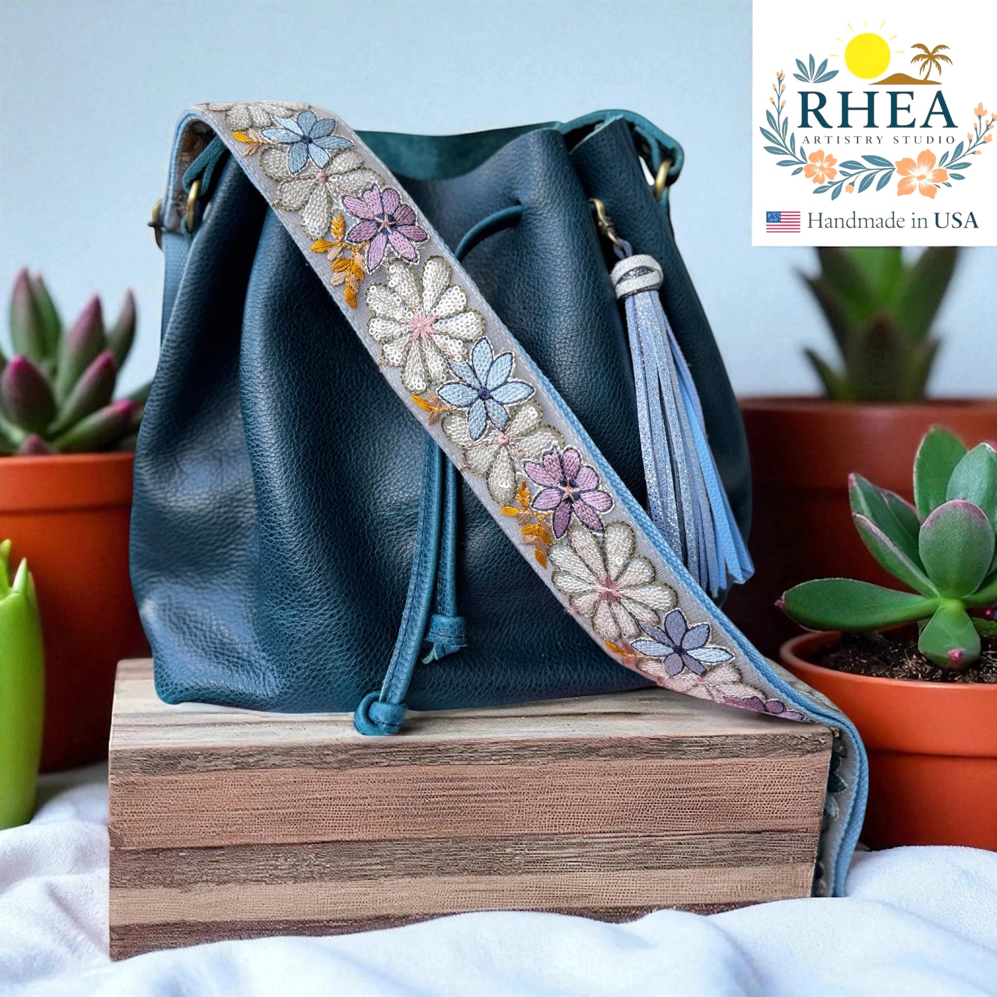 Rhea Artistry Studio 2-inch crossbody strap with embroidered daisies in pastel colors on grey lace, adjustable 30-54 inches, with brushed brass hardware.
