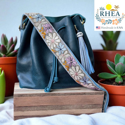 Rhea Artistry Studio 2-inch crossbody strap with embroidered daisies in pastel colors on grey lace, adjustable 30-54 inches, with brushed brass hardware.