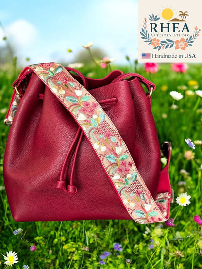 Rhea Artistry Studio red leather bucket bag with a 2-inch wide embroidered floral strap in pink, blue, and green.