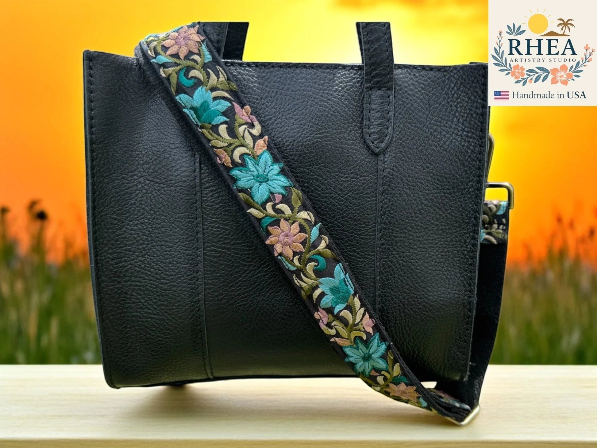 Rhea Artistry Studio black leather bag with a 1.5-inch wide adjustable crossbody strap featuring vibrant floral embroidery in teal, pink, and green.