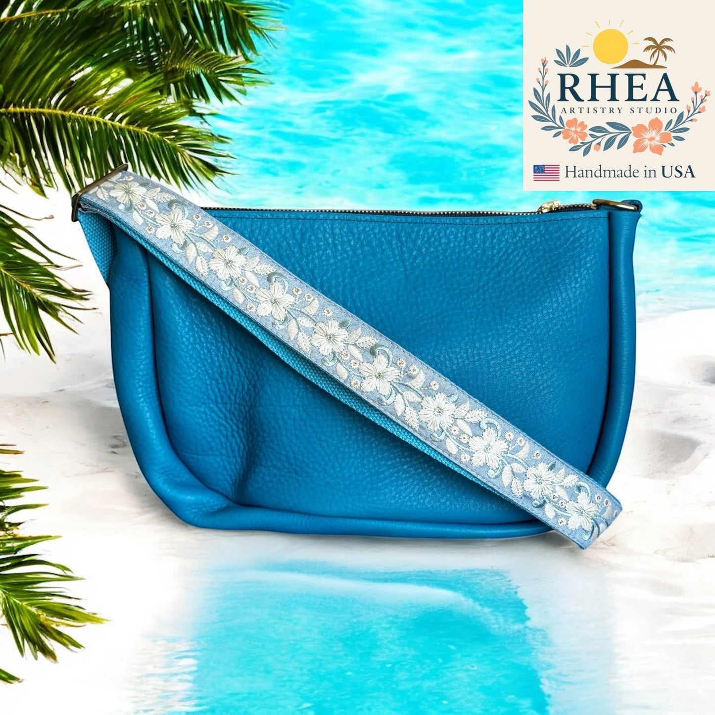 Rhea Artistry Studio blue leather crossbody bag with a 1.5-inch adjustable strap featuring white floral embroidery and brushed brass hardware.