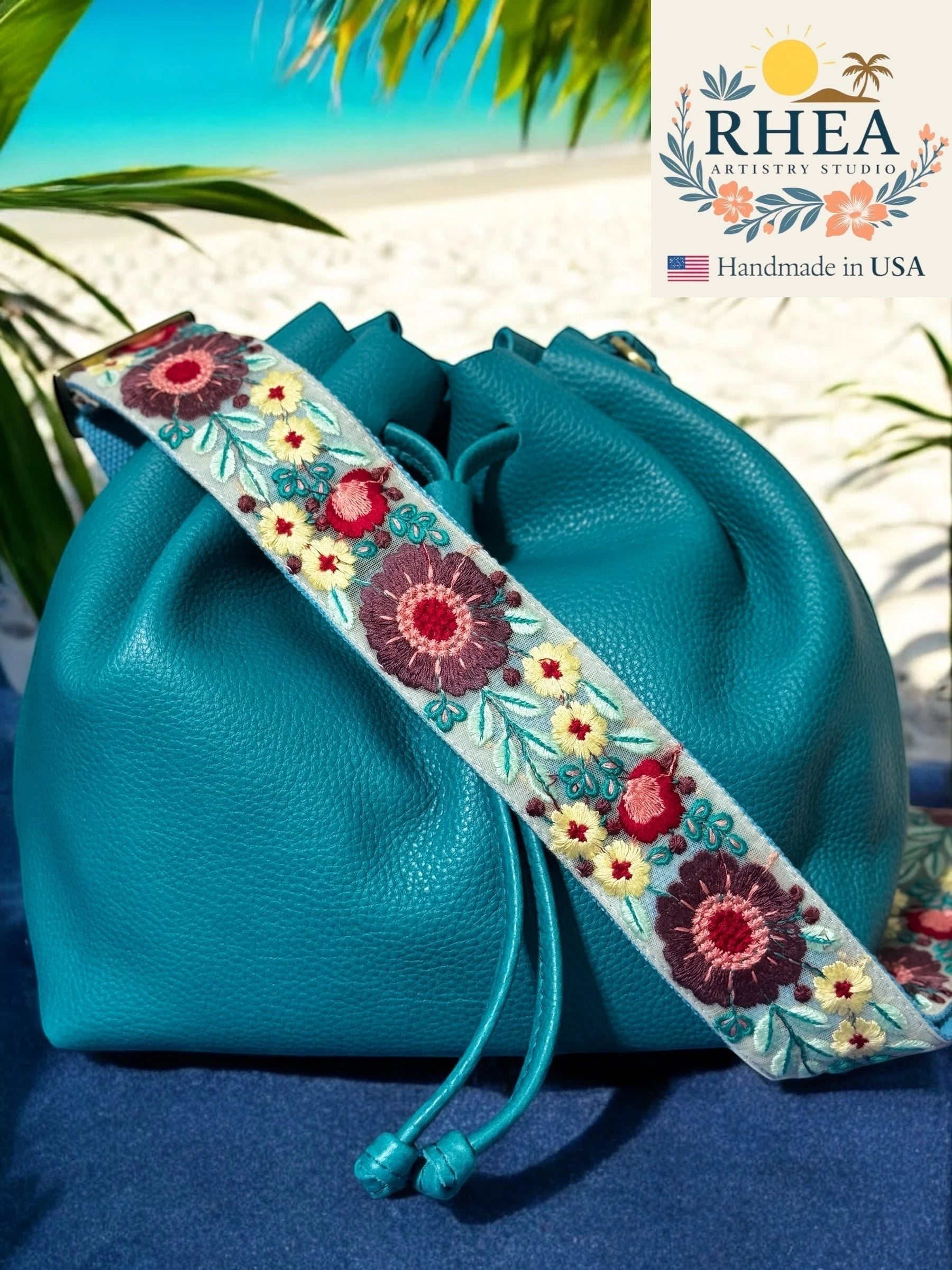 Rhea Artistry Studio 2-inch crossbody strap with embroidered floral lace in burgundy, pink, and yellow on a light blue background. Adjustable strap with brass hardware.