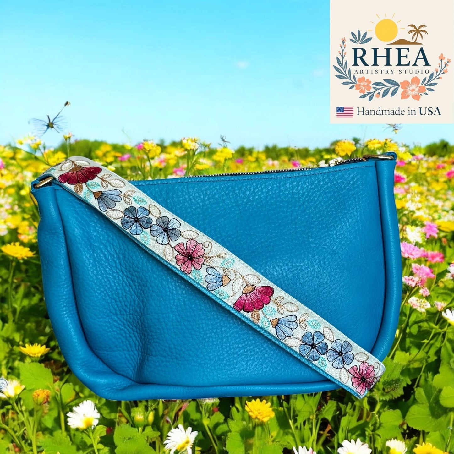 Rhea Artistry Studio blue leather crossbody bag with a 1.5-inch embroidered strap featuring pink and blue floral details, adjustable length, and brushed brass hardware.