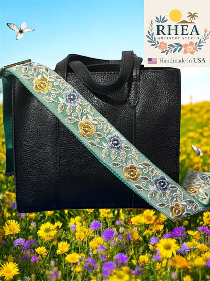 Rhea Artistry Studio 2-inch crossbody strap with embroidered gold, blue, and white florals on a light green mesh over dark webbing. Adjustable strap with brass hardware.