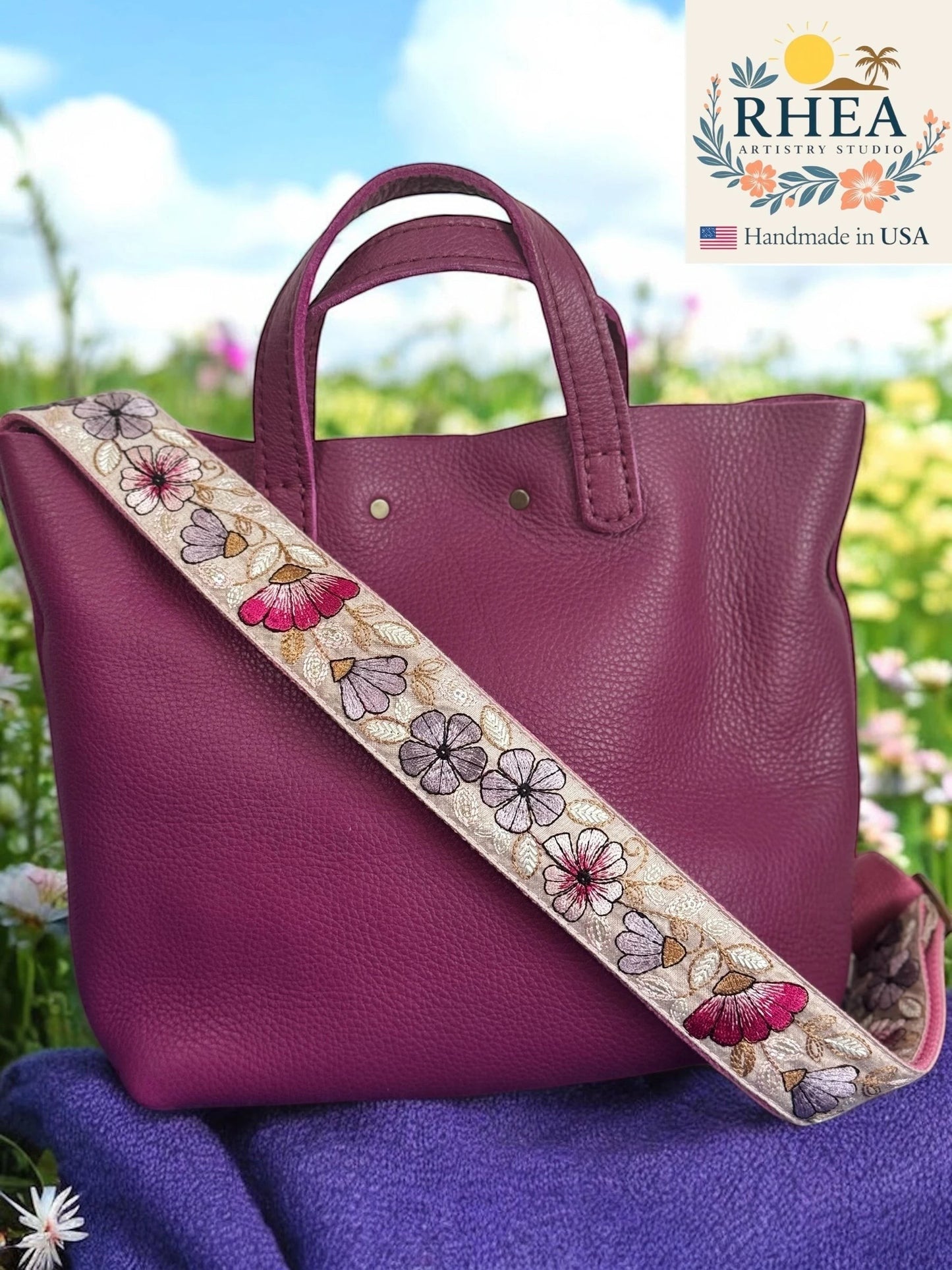 Rhea Artistry Studio 1.5-inch crossbody strap with embroidered floral pattern in pink and purple on a cream background, adjustable length with brass hardware.