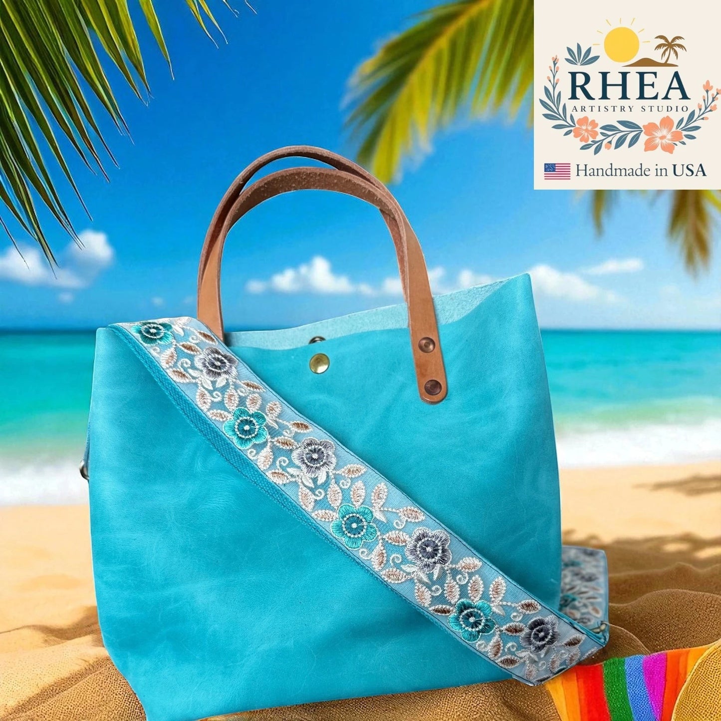 Rhea Artistry Studio 2-inch crossbody strap with embroidered blue and grey florals on a dark background, attached to a turquoise tote bag on a beach.