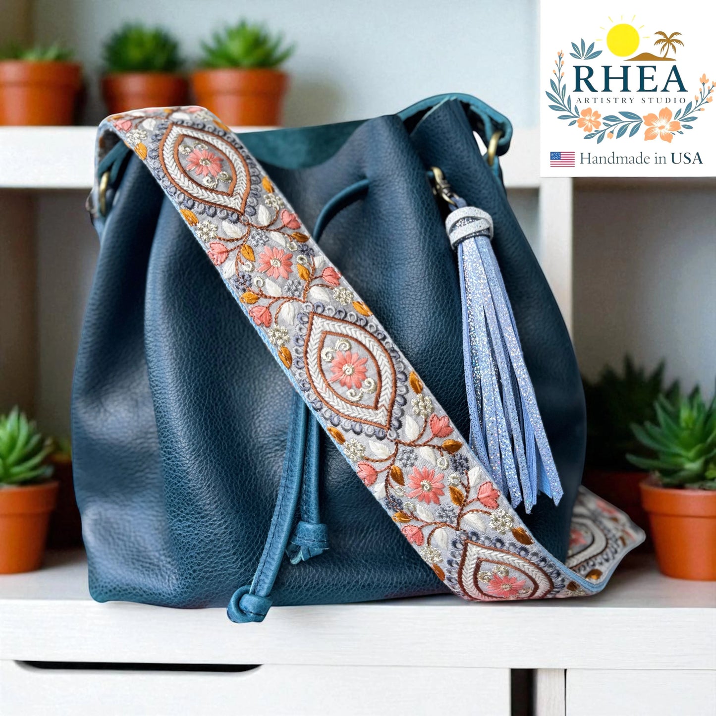 Rhea Artistry Studio 2-inch wide crossbody strap with embroidered floral panels in coral, grey, and gold on cotton webbing. Adjustable length with brushed brass hardware.
