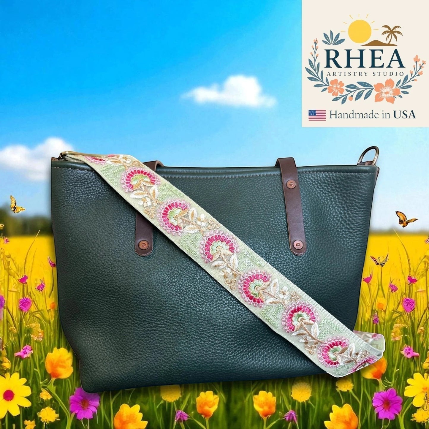 Rhea Artistry Studio 2-inch wide crossbody strap with embroidered floral lace in pink and gold on a light green background, attached to a dark green leather tote bag.