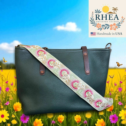 Rhea Artistry Studio 2-inch wide crossbody strap with embroidered floral lace in pink and gold on a light green background, attached to a dark green leather tote bag.