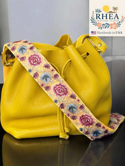 Rhea Artistry Studio 2-inch crossbody strap with yellow, pink, and blue floral embroidery on a light yellow background, attached to a yellow leather bucket bag.