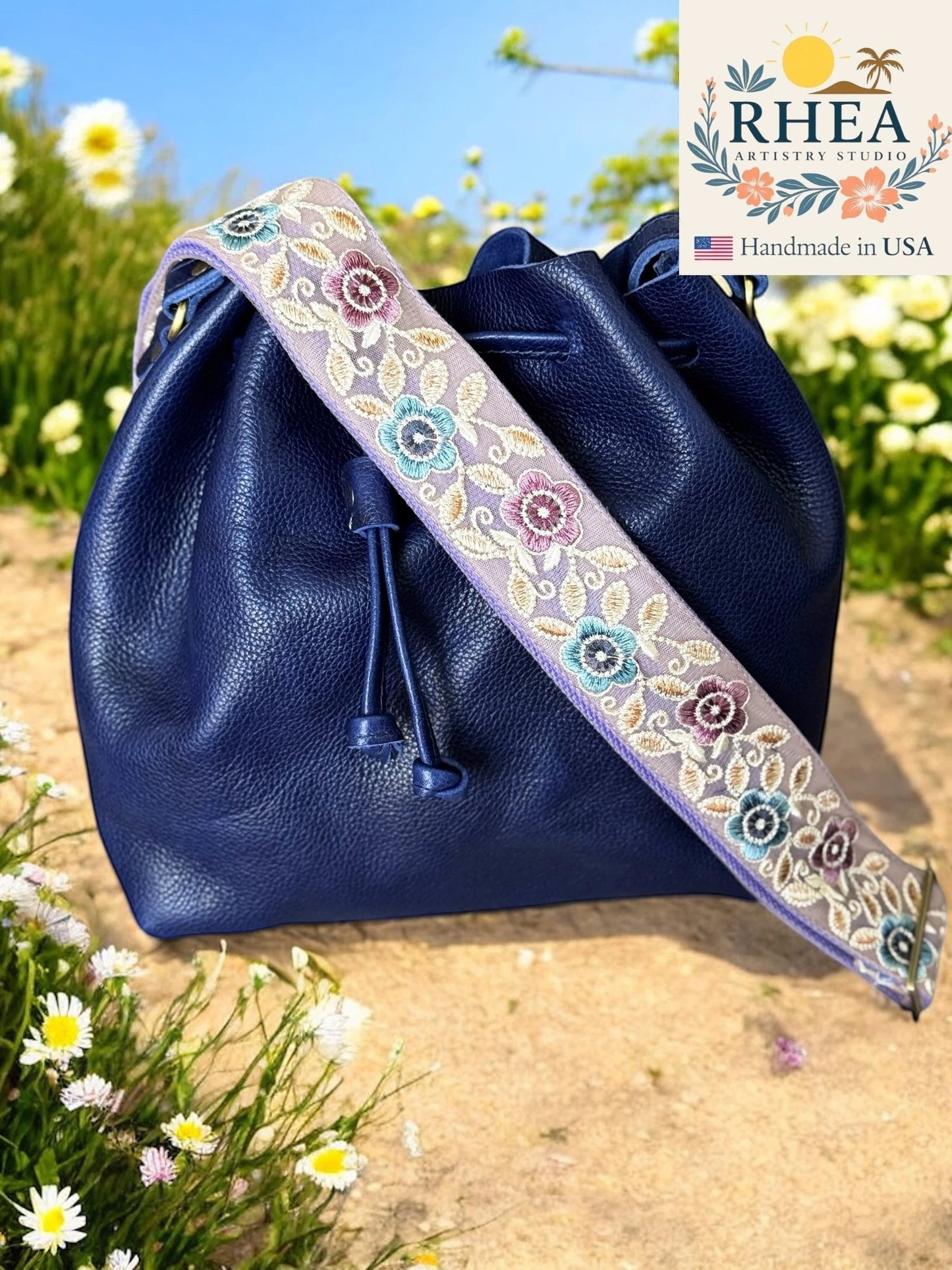 Rhea Artistry Studio 2-inch crossbody strap with embroidered pastel florals on a light purple background, attached to a navy blue leather bucket bag.
