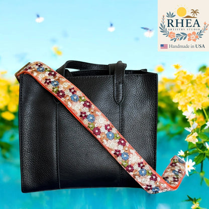 Rhea Artistry Studio embroidered crossbody strap with floral pattern in cream, blue, and burgundy on a coral edge.