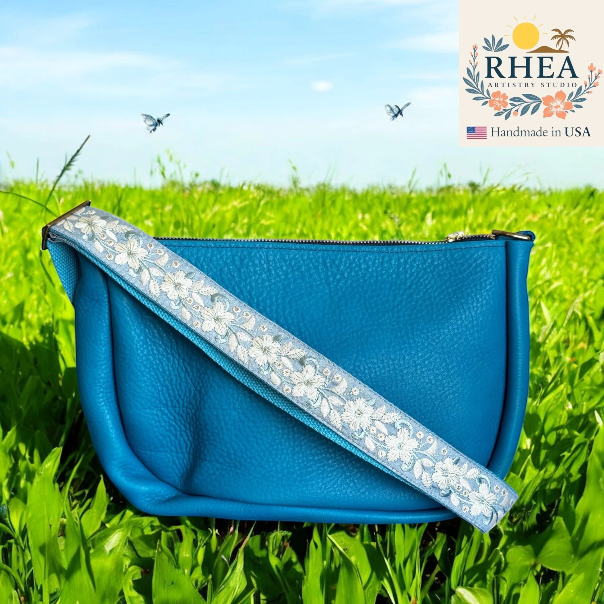 Rhea Artistry Studio blue leather crossbody bag with a 1.5-inch adjustable strap featuring white floral embroidery and brushed brass hardware.