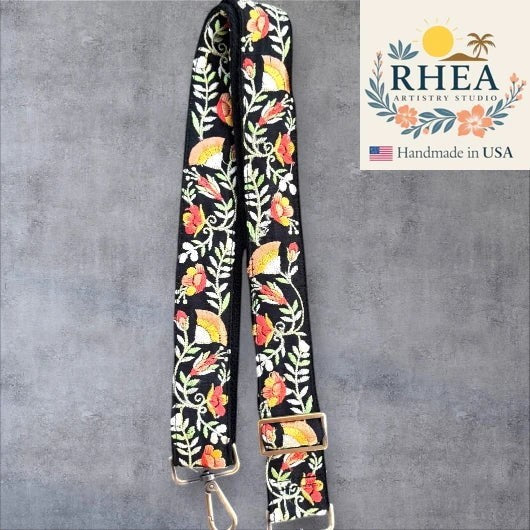 Rhea Artistry Studio black crossbody strap with embroidered floral and fan motifs in coral, yellow, and green. Adjustable length with brushed brass hardware.