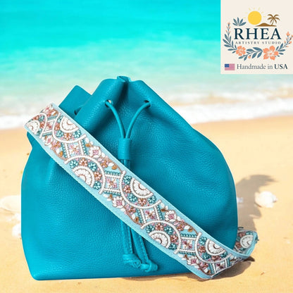 Rhea Artistry Studio 2-inch crossbody strap with embroidered floral lace in blue, pink, and white on a light blue background, attached to a teal leather bucket bag on a beach.