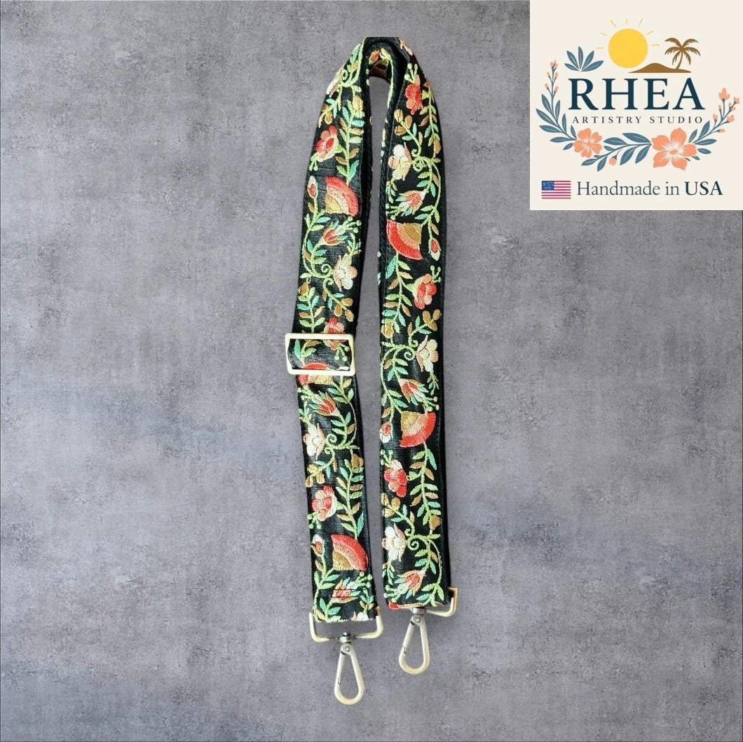Rhea Artistry Studio black crossbody strap with embroidered floral pattern in red, green, and gold. Adjustable length with brushed brass hardware.